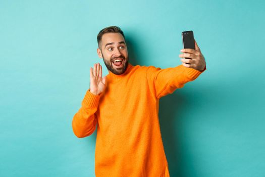 Happy young man video calling, talking online with mobile phone, saying hello to smartphone camera and waving hand friendly, standing over light blue background.