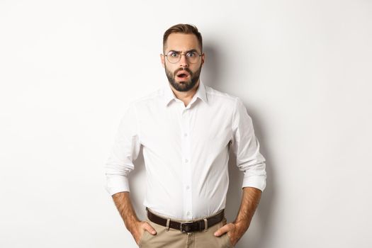 Shocked and displeased businessman in glasses, gasping and looking upset at camera, standing over white background. Copy space