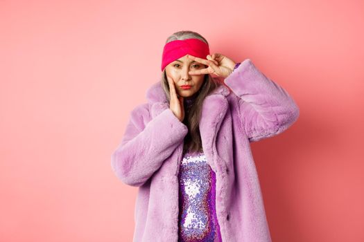 Fashion and shopping. Beautiful asian senior woman in stylish faux fur coat and headband, making peace sign on face and looking sassy and confident at camera, pink background.