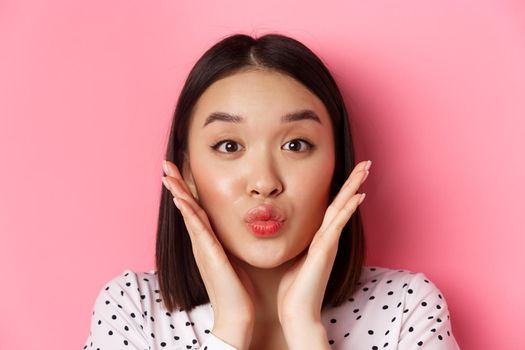 Beauty and lifestyle concept. Close-up of adorable asian woman touching face, pucker lips in kiss, standing over pink background.