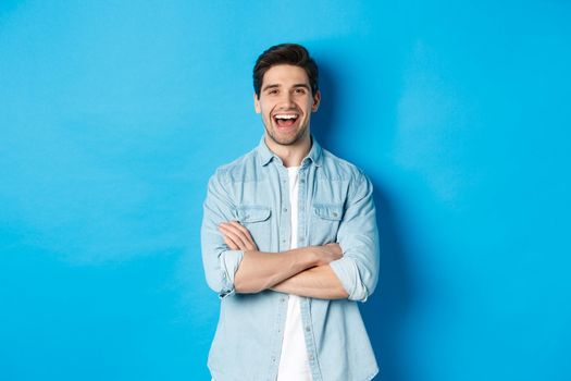 Handsome guy with beard, wearing casual outfit, laughing and looking at something funny, standing against blue background.