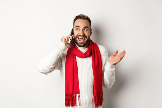 Happy bearded man wishing merry christmas on phone, calling someone and talking, standing in sweater with red scarf, white background.