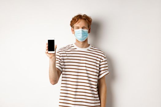 Covid-19, pandemic and social distancing concept. Handsome young man with face mask, showing black smartphone screen and smiling, standing over white background.