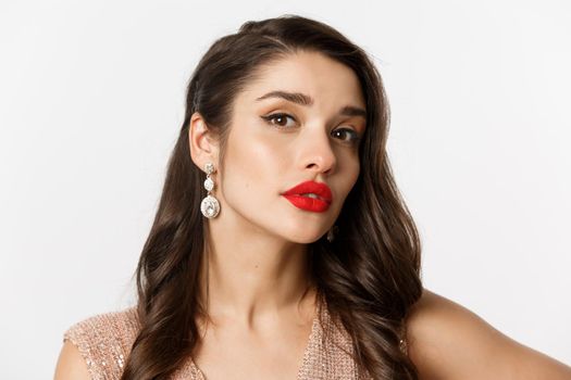 Concept of New Year celebration and winter holidays. Close-up of sensual and beautiful woman with red lips, looking confident at camera, wearing luxury earrings, white background.