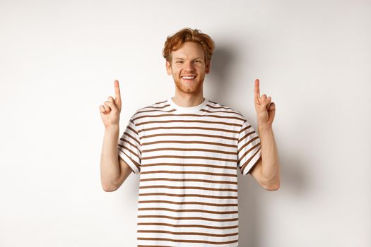Smiling young male student with red hair showing logo, pointing fingers up and looking happy, standing over white background.