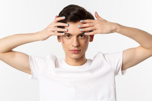 People, lgbtq and beauty concept. Close-up of beautiful queer man touching face with fingers with blue nail polish, standing over white background.