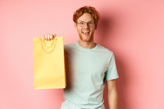 Cheerful redhead man in t-shirt, showing shopping bag and smiling, standing over pink background.