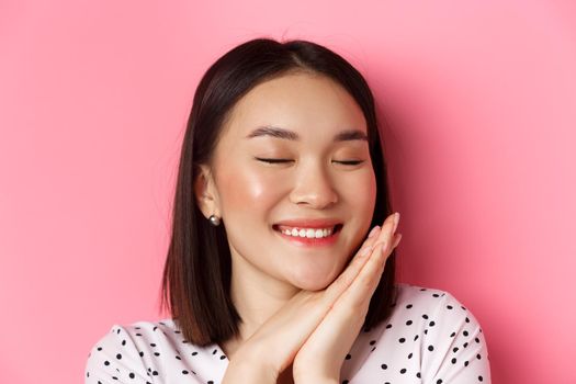 Beauty and skin care concept. Headshot of adorable and dreamy asian woman close eyes, smiling nostalgic, standing against pink background.