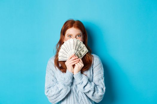Excited redhead woman covering face with money, holding dollars and staring at camera happy, standing over blue background.