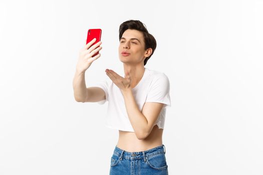 Beautiful gay man in crop top sending air kiss at phone camera, taking selfie or video chat on smartphone, standing over white background.