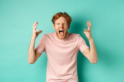 Angry young redhead man screaming distressed, shaking hands and looking outraged, standing over turquoise background. Copy space