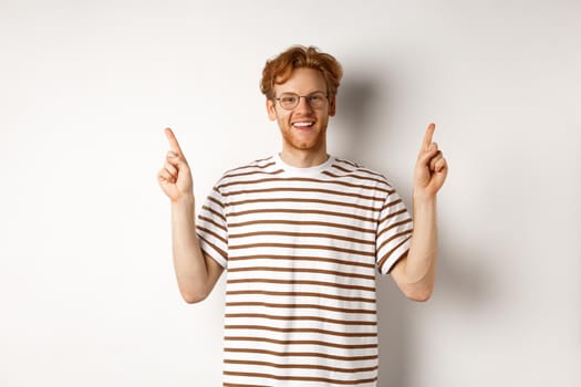 Funny young redhead guy in nerdy glasses pointing fingers sideways, showing two promo offers, standing happy over white background.