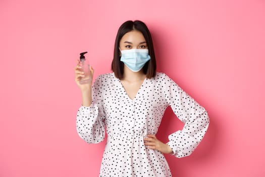 Covid-19, pandemic and lifestyle concept. Beautiful korean woman in dress and medical mask showing hand sanitizer, recommending antiseptic, standing over pink background.