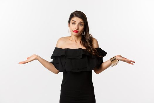 Fashion and beauty. Clueless attractive woman in black dress dont know, shrugging and smirking unaware, standing indecisive over white background.