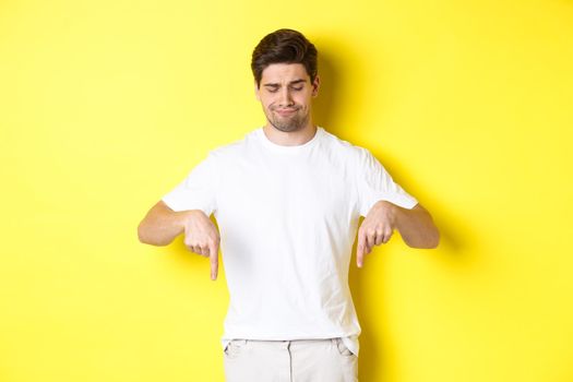 Skeptical young man in white t-shirt, pointing and looking down upset, disapprove and dislike product, standing over yellow background.