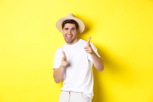 Concept of tourism and vacation. Happy cool guy pointing fingers at you, standing over yellow background.