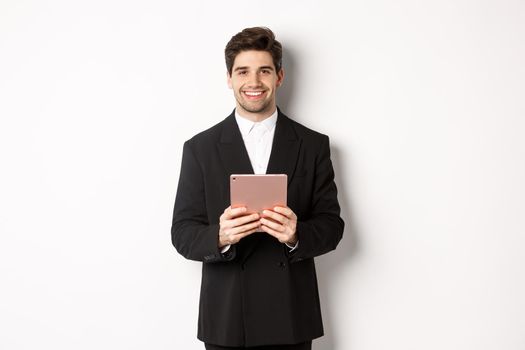 Image of handsome businessman in trendy suit, holding digital tablet and smiling, standing against white background.