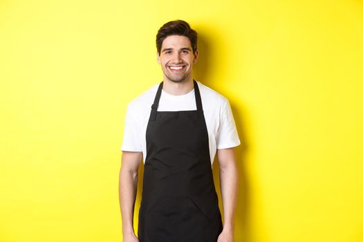 Young man waiter in black apron, smiling, working in store or coffee shop, standing against yellow background.