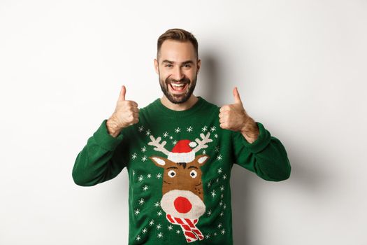 Christmas, holidays and celebration. Happy young man enjoying New Year party, showing thumbs up in approval, like something good, standing over white background.