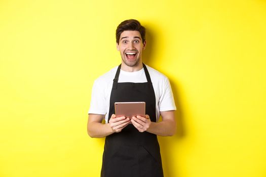Happy salesman in black apron, holding digital tablet and looking surprised, standing against yellow background.