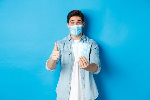 Concept of coronavirus, quarantine and social distancing. Young man showing medical masks and thumb up, standing against blue background.