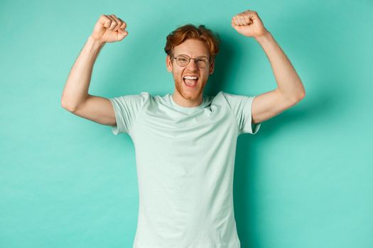 Image of young redhead man feeling like champion, raising hands up in fist pump gesture and shouting yes with joy, winning prize, triumphing of success, standing over mint background.