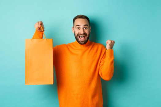 Happy man holding orange shopping bag and rejoicing, got discount and celebrating, standing over turquoise background.