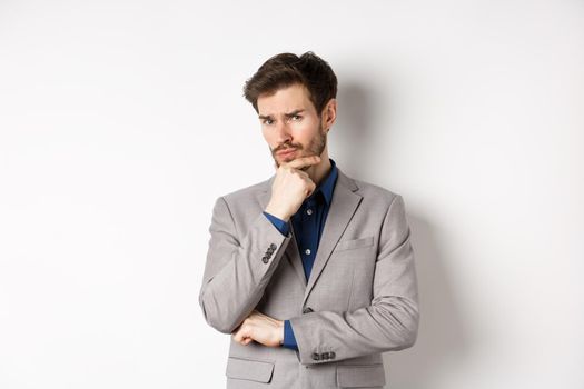Hesitant young businessman in suit looking skeptical at camera, thinking and making decision, standing against white background.