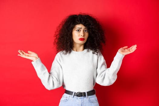 Who knows. Clueless young woman with curly hair, shrugging and looking confused, standing in casual clothes on red background.