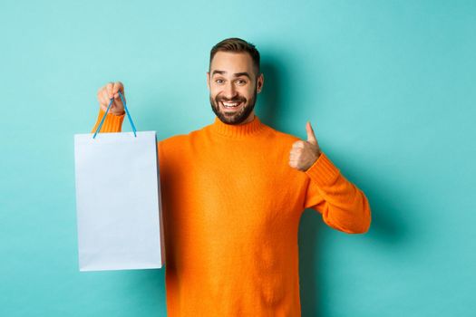 Handsome smiling man showing thumbs-up and shopping bag, recommending store, standing over blue background. Copy space