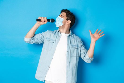 Handsome guy in medical mask singing in microphone, standing over blue background.
