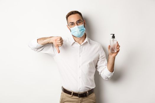 Covid-19, social distancing and quarantine concept. Office worker in medical mask displeased, showing hand sanitizer and thumb down, standing over white background.