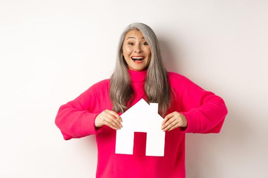 Real estate. Excited asian senior woman looking happy and lucky, winning apartment, showing paper house model and smiling, standing over white background.