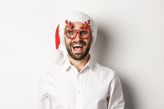 Close-up of excited bearded man in christmas glasses and santa hat looking amazed at promo offer, winter holidays advertisement concept, white background.