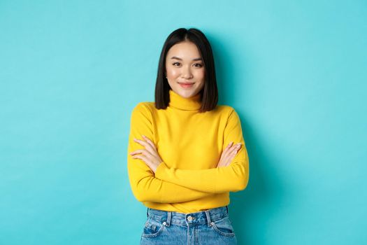 Attractive asian woman with short dark hair, cross arms on chest and smiling confident, standing over blue background.