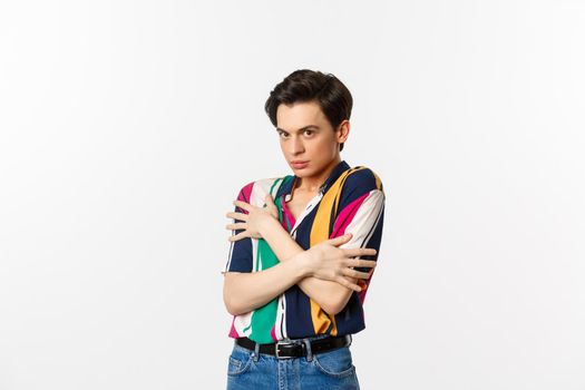 Image of timid gay man cringe and looking displeased at camera, hugging himself defensive, standing over white background.