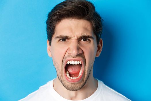 Head shot of angry man shouting and looking with hatred, standing mad against blue background.