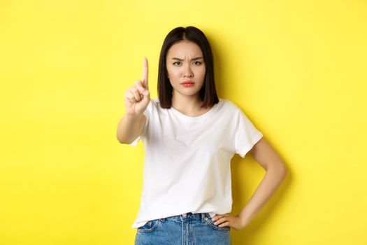 Confident and serious woman tell no, showing extended finger to stop and prohibit something bad, frowning and looking at camera self-assured, standing over yellow background.