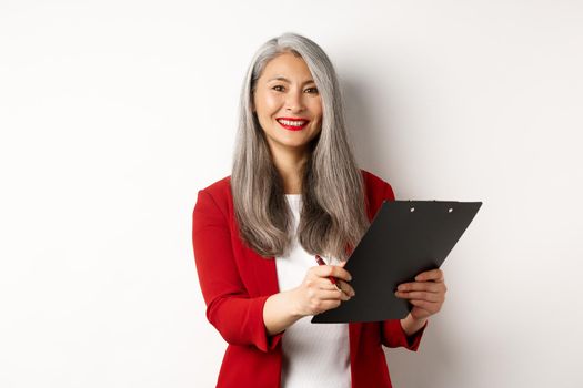 Elegant asian senior businesswoman working with documents, holding pen and clipboard, signing contract and smiling, standing over white background.