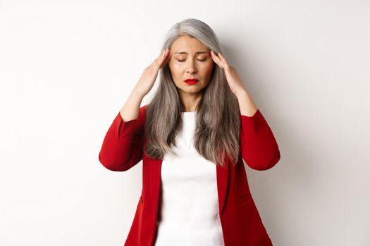 Distressed asian businesswoman trying to calm down, massaging temples on head to soothe headache, standing over white background.