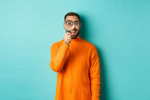 Cheerful man searching for something, looking through magnifying glass and smiling happy, standing over blue background.