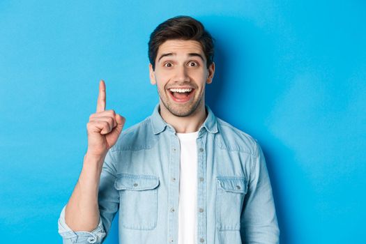 Close-up of handsome bearded guy smiling, showing finger number one, standing over blue background.