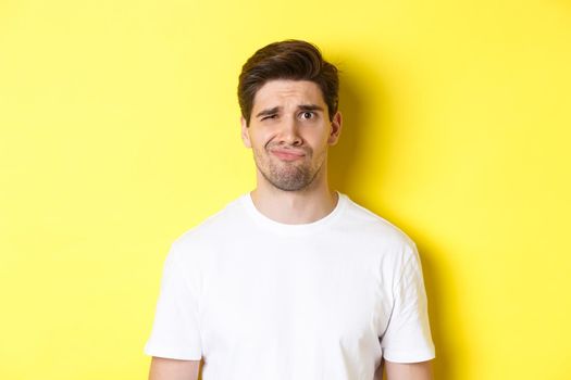 Close-up of dispelased young man in white t-shirt looking doubtful, grimacing unsatisfied, standing over yellow background.