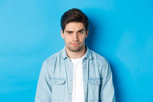 Close-up of suspicious and serious guy raising one eyebrow, looking at camera, standing against blue background.