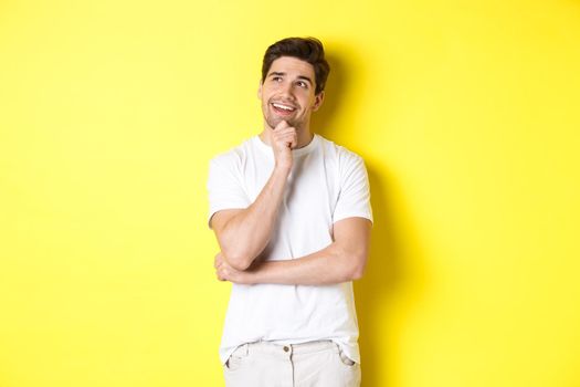 Portrait of smiling man looking thoughtful at upper left corner, choosing something, have an idea, standing against yellow background.