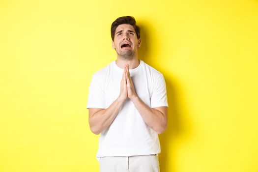 Desperate man pleading God, holding hands in pray and looking up distressed, standing over yellow background. Copy space