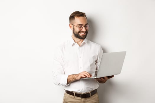 Business. Handsome businessman working on laptop, answering messages and smiling, standing over white background.