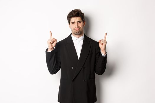 Portrait of reluctant and sad handsome man, complaining and pointing fingers up at something bad, looking jealous, standing over white background.