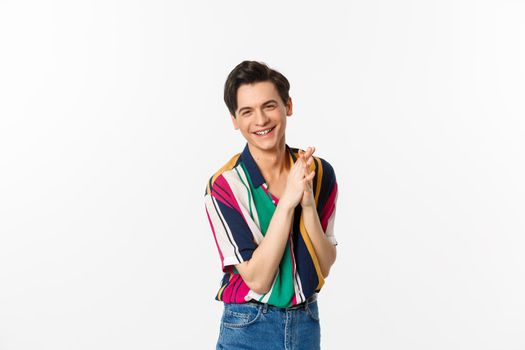 Attractive young slim man relish something good, rubbing hands and smiling satisfied, standing over white background. Copy space