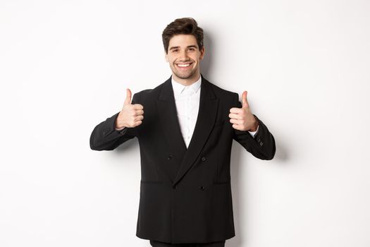 Concept of new year party, celebration and lifestyle. Image of attractive happy businessman in formal suit, showing thumbs-up and smiling, like and approve, standing over white background.